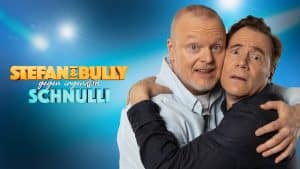 Stefan Raab und Michael Bully Herbig umarmen sich humorvoll vor einem blauen Hintergrund mit dem Schriftzug „Stefan & Bully gegen irgendson Schnulli“. Der Titel der RTL-Show ist in auffälligen Farben gestaltet.