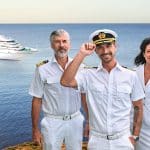 Staff-Kapitän Martin Grimm (Daniel Morgenroth), Kapitän Max Parger (Florian Silbereisen) und Hoteldirektorin Hanna Liebhold (Barbara Wussow) in weißer Uniform stehen vor einem malerischen Hintergrund mit der Amadea, dem Traumschiff, das auf dem ruhigen blauen Meer zu sehen ist.