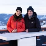 Lena Kesting (links) trägt eine rote Steppjacke und eine schwarze Mütze, während sie neben Severin Freund (rechts) in schwarzer Jacke und schwarzer Mütze an einem Moderationstisch steht. Beide lächeln in die Kamera. Im Hintergrund ist ein verschneites Bergpanorama und das Skistadion von Oberstdorf zu sehen.