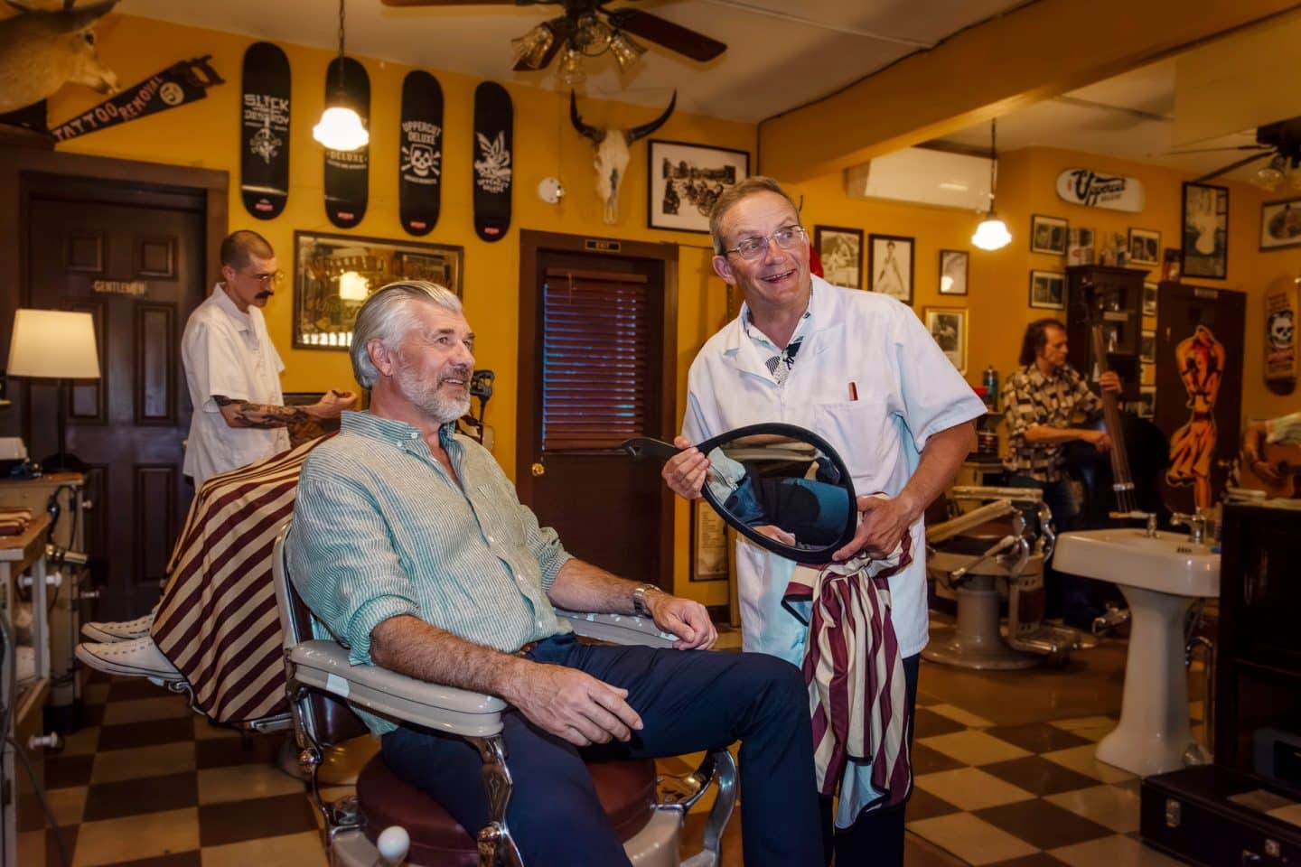 "Das Traumschiff: Hudson Valley": Im Barbershop: Lachend sitzt Martin Grimm (Daniel Morgenroth) auf dem Friseurstuhl. Neben ihm steht, ebenfalls lachend, der Barbier (Wigald Boning) und hält einen Spiegel und ein Handtuch in den Händen. Im Hintergrund werden weitere Kunden bedient (Komparsen).