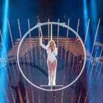 Helene Fischer steht in einem schwebenden Ring auf der Bühne, während sie singt. Sie trägt ein weißes, glamouröses Outfit, und der Hintergrund ist mit strahlenden Lichtbalken und goldenen Akzenten spektakulär beleuchtet.
