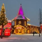 Ein weihnachtlich dekoriertes Holzgebäude mit einem spitzen Dach, beleuchtet in warmen Farben und umgeben von schneebedeckten Bäumen. Ein großer Weihnachtsbaum mit leuchtenden Lichtern steht links vom Gebäude, und mehrere beleuchtete Schilder weisen auf eine Weihnachtsattraktion hin. Die Szenerie ist in winterlicher Atmosphäre gehalten.