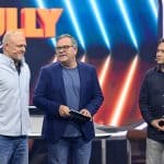 Stefan Raab, Moderator Elton und Michael 'Bully' Herbig stehen gemeinsam auf der Bühne der Show 'Stefan & Bully gegen irgendson Schnulli', vor einem leuchtenden Hintergrund mit dem Show-Logo.