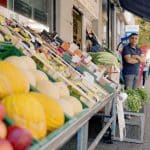 Ein türkischer Supermarkt mit frischen Obst- und Gemüseauslagen vor dem Geschäft. Ein Mann steht lächelnd mit verschränkten Armen daneben, während Passanten die Straße entlanggehen.