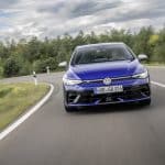 Ein blauer Volkswagen Golf R fährt dynamisch auf einer kurvigen Landstraße, umgeben von grüner Vegetation und bewölktem Himmel.