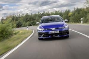Ein blauer Volkswagen Golf R fährt dynamisch auf einer kurvigen Landstraße, umgeben von grüner Vegetation und bewölktem Himmel.