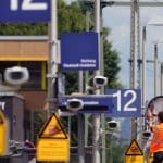 Ein Arbeiter mit orangefarbener Warnweste repariert eine Uhr an einem Bahnsteig während der Riedbahnsanierung. Im Vordergrund sind Warnschilder und blaue Bahnsteignummern mit der Aufschrift „12“ zu sehen. Die Umgebung zeigt typische Bahninfrastruktur, inklusive Überwachungskameras, Lampen und Fahrleitungen.