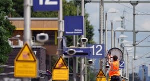 Ein Arbeiter mit orangefarbener Warnweste repariert eine Uhr an einem Bahnsteig während der Riedbahnsanierung. Im Vordergrund sind Warnschilder und blaue Bahnsteignummern mit der Aufschrift „12“ zu sehen. Die Umgebung zeigt typische Bahninfrastruktur, inklusive Überwachungskameras, Lampen und Fahrleitungen.