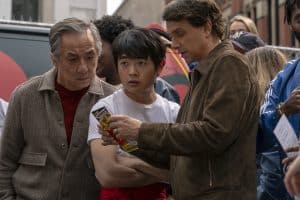 Jackie Chan, Ben Wang und Ralph Macchio in einer Szene aus ‚Karate Kid: Legends‘ – gemeinsames Studium eines Flyers in einer belebten Umgebung.