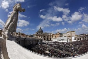 Ein Blick über den Petersplatz im Vatikan, mit der imposanten Kuppel des Petersdoms im Hintergrund. Eine große Menschenmenge füllt den Platz, während eine Statue im Vordergrund majestätisch auf die Szene blickt. Der Himmel ist klar mit einigen Wolken.