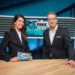 Linda Zervakis und ein männlicher Kollege stehen lächelnd in einem modernen Nachrichtenstudio. Auf dem Bildschirm im Hintergrund ist das Logo „Fake News – Alles erstunken und erlogen“ zu sehen. Beide tragen elegante Anzüge und wirken professionell.
