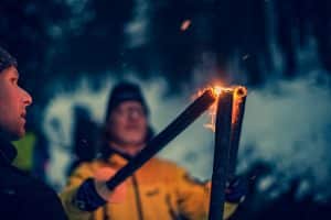Menschen entzünden Fackeln in einer verschneiten Winterlandschaft, umgeben von dunklen Bäumen und einer stimmungsvollen Atmosphäre.
