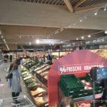 Innenansicht eines modernen Supermarkts mit Holzbalkendecke. Kunden stöbern in den Gemüse- und Obstregalen. Im Vordergrund ist ein Angebotstisch mit der Aufschrift '% Frische Kracher %' zu sehen.