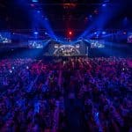 Die Europa-Park Arena während der Darts-Gala: Eine voll besetzte Halle mit stimmungsvoller Beleuchtung, zahlreichen Fans an Tischen und einer Bühne, auf der die Darts-Spieler im Rampenlicht stehen. Großbildschirme zeigen Live-Bilder der Spieler.