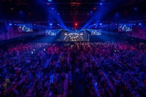 Die Europa-Park Arena während der Darts-Gala: Eine voll besetzte Halle mit stimmungsvoller Beleuchtung, zahlreichen Fans an Tischen und einer Bühne, auf der die Darts-Spieler im Rampenlicht stehen. Großbildschirme zeigen Live-Bilder der Spieler.