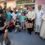 Eine Gruppe von Kindern, Jugendlichen, Krankenhauspersonal und Gästen posiert in einem Spielzimmer der Kinderklinik Ortenau. In der Mitte stehen Ed Euromaus, maskottchenartig als Weihnachtsfigur gekleidet, sowie eine winterlich gekleidete Prinzessin. Mauritia Mack hält einen Korb mit Geschenken. Ein Junge im Rollstuhl mit Gipsarm sitzt im Vordergrund, umgeben von lächelnden Kindern und Erwachsenen. Im Hintergrund ist die farbenfrohe Schrift "SPIELZIMMER" zu sehen.
