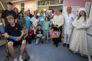 Eine Gruppe von Kindern, Jugendlichen, Krankenhauspersonal und Gästen posiert in einem Spielzimmer der Kinderklinik Ortenau. In der Mitte stehen Ed Euromaus, maskottchenartig als Weihnachtsfigur gekleidet, sowie eine winterlich gekleidete Prinzessin. Mauritia Mack hält einen Korb mit Geschenken. Ein Junge im Rollstuhl mit Gipsarm sitzt im Vordergrund, umgeben von lächelnden Kindern und Erwachsenen. Im Hintergrund ist die farbenfrohe Schrift "SPIELZIMMER" zu sehen.