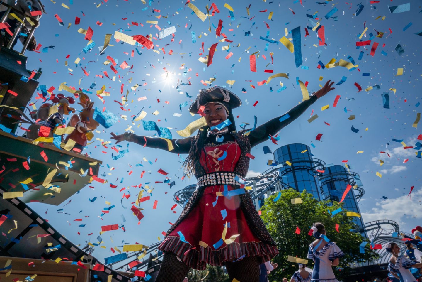 Eine fröhliche Frau im Piratenkostüm steht mit ausgebreiteten Armen unter einem Konfettiregen. Im Hintergrund ist die markante Glasfassade der Achterbahn „Euro-Mir“ im Europa-Park zu sehen. Die Szene ist lebendig, mit blauem Himmel und strahlendem Sonnenschein.