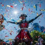 Eine fröhliche Frau im Piratenkostüm steht mit ausgebreiteten Armen unter einem Konfettiregen. Im Hintergrund ist die markante Glasfassade der Achterbahn „Euro-Mir“ im Europa-Park zu sehen. Die Szene ist lebendig, mit blauem Himmel und strahlendem Sonnenschein.