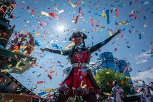 Eine fröhliche Frau im Piratenkostüm steht mit ausgebreiteten Armen unter einem Konfettiregen. Im Hintergrund ist die markante Glasfassade der Achterbahn „Euro-Mir“ im Europa-Park zu sehen. Die Szene ist lebendig, mit blauem Himmel und strahlendem Sonnenschein.