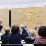 Abgeordnete des Landtags von Baden-Württemberg heben in einer Plenarsitzung die Hände zur Abstimmung. Im Hintergrund ist das Landeswappen Baden-Württembergs zu sehen, während Redner an einem Podium stehen.