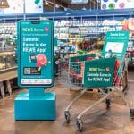 Eine Verkaufsfläche in einem REWE-Markt mit einem großen digitalen Display, das das neue REWE Bonusprogramm bewirbt. Auf dem Display steht „Sammle Euros in der REWE App!“. Daneben ein Einkaufswagen, ebenfalls mit Werbehinweisen für das Bonusprogramm, der mit Lebensmitteln gefüllt ist. Im Hintergrund sind Regale mit frischen Lebensmitteln sichtbar.