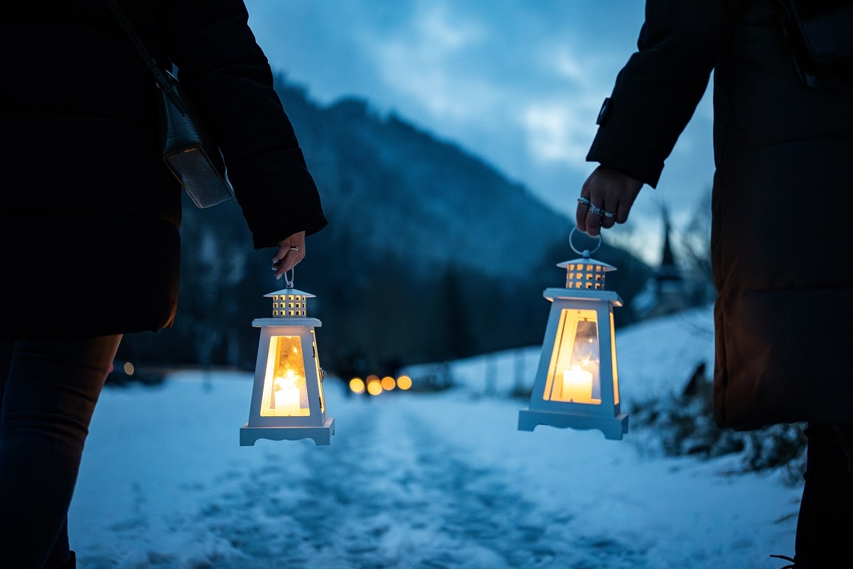 Zwei Personen tragen Laternen durch eine verschneite Winterlandschaft bei Einbruch der Dämmerung.

