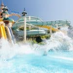 "Ein farbenfroher Wasserpark mit mehreren Wasserrutschen, darunter spiralförmige und gerade Rutschen, die in ein großes, sprudelndes Becken münden. Im Hintergrund sind türmchenartige Strukturen und ein Schild mit der Aufschrift 'Aqua Fantasy' zu sehen, unter klarem, blauem Himmel.
