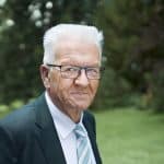 Winfried Kretschmann, Ministerpräsident von Baden-Württemberg, in einem Anzug mit Krawatte. Im Hintergrund sind Bäume und Sträucher in einer grünen, natürlichen Umgebung zu sehen.
