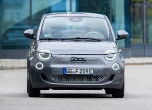 Ein grauer Fiat 500 Elektro steht frontal auf einer Straße vor einem modernen Gebäude mit Glasfassade. Das Nummernschild trägt die Kennung „GG F 259E“, und der Wagen zeigt das Fiat-Logo auf der Vorderseite. Die runden LED-Scheinwerfer sind eingeschaltet.