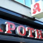 Außenansicht einer Apotheke mit leuchtenden roten Buchstaben und dem Apotheken-Logo auf einem Würfelschild an der Fassade