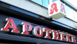 Außenansicht einer Apotheke mit leuchtenden roten Buchstaben und dem Apotheken-Logo auf einem Würfelschild an der Fassade