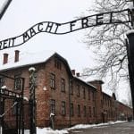 Das Eingangstor des ehemaligen Konzentrationslagers Auschwitz mit der Aufschrift "Arbeit macht frei" im Winter. Schnee bedeckt das Gelände und die Gebäude aus rotem Ziegelstein, während kahle Bäume den Hintergrund säumen.