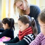 Eine Lehrerin beugt sich über eine Gruppe von Grundschulkindern, die konzentriert an ihren Tischen arbeiten. Die Kinder malen und schreiben mit farbigen Stiften, während die Lehrerin unterstützend zur Seite steht. Der Raum ist hell und freundlich gestaltet.