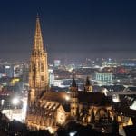 Nachtaufnahme des Freiburger Münsters, einer beleuchteten gotischen Kathedrale, umgeben von der Stadt Freiburg. Im Hintergrund erstreckt sich die Skyline der Stadt, die ebenfalls von Lichtern erhellt wird, mit dem Schwarzwald in der Ferne.