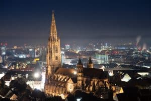Nachtaufnahme des Freiburger Münsters, einer beleuchteten gotischen Kathedrale, umgeben von der Stadt Freiburg. Im Hintergrund erstreckt sich die Skyline der Stadt, die ebenfalls von Lichtern erhellt wird, mit dem Schwarzwald in der Ferne.