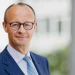 Friedrich Merz, Vorsitzender der CDU, steht in einem dunkelblauen Anzug und einer gemusterten Krawatte in einem modernen, lichtdurchfluteten Gebäude. Er trägt eine Brille und hat einen freundlichen, selbstbewussten Gesichtsausdruck. Der Hintergrund ist unscharf, wodurch der Fokus auf ihm liegt.