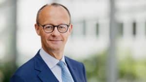 Friedrich Merz, Vorsitzender der CDU, steht in einem dunkelblauen Anzug und einer gemusterten Krawatte in einem modernen, lichtdurchfluteten Gebäude. Er trägt eine Brille und hat einen freundlichen, selbstbewussten Gesichtsausdruck. Der Hintergrund ist unscharf, wodurch der Fokus auf ihm liegt.