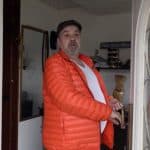 Frank, ein Mann mittleren Alters mit grauem Bart und Mütze, steht in der offenen Haustür. Er trägt eine auffällige orangefarbene Steppjacke und blickt überrascht oder fragend in Richtung der Kamera. Im Hintergrund ist ein Flur mit Dekorationen und einem Spiegel sichtbar.