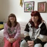 Petra sitzt mit ihrer Tochter Selina auf einem Sofa. Beide lächeln, während Petra von ihrer neuen Liebe erzählt. Im Hintergrund sind persönliche Dekorationen und Bilder an der Wand zu sehen, die das Wohnzimmer gemütlich wirken lassen.