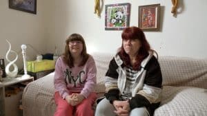 Petra sitzt mit ihrer Tochter Selina auf einem Sofa. Beide lächeln, während Petra von ihrer neuen Liebe erzählt. Im Hintergrund sind persönliche Dekorationen und Bilder an der Wand zu sehen, die das Wohnzimmer gemütlich wirken lassen.