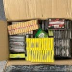 Offener Karton mit mehreren Packungen Feuerwerkskörper, darunter Böller in gelben, schwarzen und roten Verpackungen, die eng gestapelt sind.