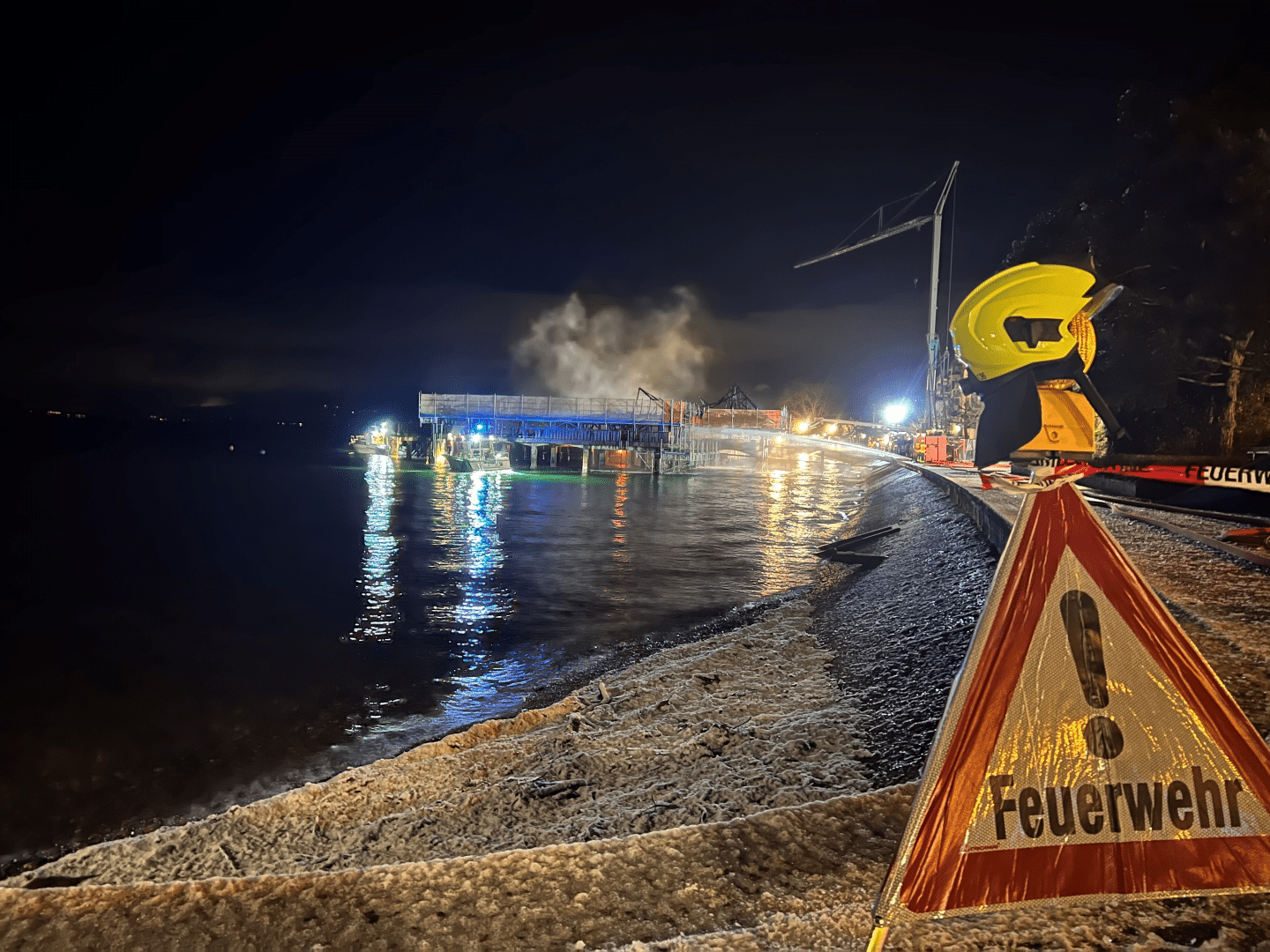 Die Rorschacher Badhütte am Schweizer Bodenseeufer nach dem Brand in der Nacht: Das hölzerne Stelzengebäude raucht noch leicht, während Feuerwehrleute am Ufer ihre Löscharbeiten beenden.

