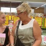 Loredana und Silvia Wollny stehen lächelnd in einem Supermarkt. Loredana hält eine pinkfarbene Box in der Hand, während Silvia, die eine Brille und ein grau-weißes Kleid trägt, sie freundlich ansieht. Im Hintergrund sind Preisschilder und Supermarktregale zu sehen.