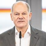 Bundeskanzler Olaf Scholz während einer Rede. Er trägt ein graues Sakko und ein weißes Hemd, spricht in ein Mikrofon und steht vor einem hellen Hintergrund.
