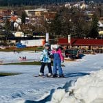 Zwei Kinder stehen auf Skiern auf einer schneebedeckten Piste in der Max Wild Arena in Isny, mit Blick auf die Stadt im Hintergrund. Die Szenerie zeigt einen sonnigen Wintertag, während im Hintergrund der Schlepplift und die gemütliche Einkehrhütte zu sehen sind.