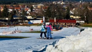 Zwei Kinder stehen auf Skiern auf einer schneebedeckten Piste in der Max Wild Arena in Isny, mit Blick auf die Stadt im Hintergrund. Die Szenerie zeigt einen sonnigen Wintertag, während im Hintergrund der Schlepplift und die gemütliche Einkehrhütte zu sehen sind.