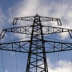 Ein deutscher Strommast aus Stahl mit dreifacher Quertraverse, der sich vor einem bewölkten Himmel erhebt. Die hochgespannten Stromleitungen sind deutlich sichtbar und verlaufen in verschiedene Richtungen. Der Mast zeigt eine robuste Konstruktion und dominiert die Szenerie.