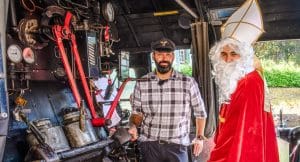 Ein Lokführer in traditioneller Uniform steht neben einer als Nikolaus verkleideten Person im Führerstand einer historischen Dampflok. Die Szene spielt im Rahmen der Sonderfahrt des Dampfzugs zum Weihnachtsmarkt in Ottenhöfen. Im Hintergrund sind technische Details der Dampflok und eine grüne Außenlandschaft erkennbar.