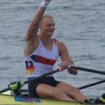 Oliver Zeidler in Paris 2024, deutscher Ruderer, sitzt in einem Einer-Ruderboot auf dem Wasser und winkt mit der rechten Hand. Er trägt ein weißes Deutschland-Trikot mit Bundesadler-Logo, während er mit der linken Hand ein Ruder hält.